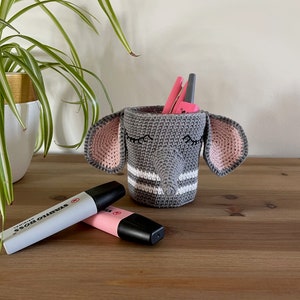 Peut inclure: Un porte-crayons en forme d'éléphant crocheté gris avec des oreilles roses et des rayures blanches. Le porte-crayons est rempli de stylos et de crayons. Deux surligneurs roses et gris sont sur une surface en bois.