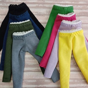 Può includere: Un set di otto paia di leggings per bambole in vari colori, tra cui nero, blu, verde, rosa, viola e giallo. Ogni paio ha un bordo in pizzo bianco in alto.
