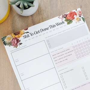 May include: A white weekly planner with a floral design and the text "Shit To Get Done This Week". The planner includes sections for daily schedule, top priorities, daily habit tracker, and shopping lists.