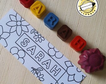 May include: A set of colorful crayons shaped like the letters S, A, R, A, H. The crayons are arranged in a line on a white surface with a floral design. The crayons are from Crayon Allsorts.