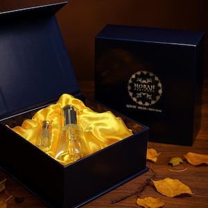 May include: A navy blue gift box with a gold logo and the text "NORAH PERFUMES". Inside, two perfume bottles rest on a bed of yellow satin. The box is open, revealing the bottles and satin. The second box is closed.