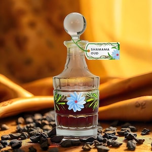 Puede incluir: Un frasco de perfume de vidrio transparente con un tap&oacute;n redondo de vidrio. La botella est&aacute; llena de un l&iacute;quido oscuro y decorada con una flor azul y hojas verdes. Una etiqueta dice "SHAMAMA OUD".