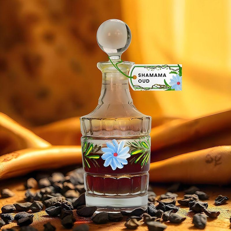 Puede incluir: Un frasco de perfume de vidrio transparente con un tap&oacute;n redondo de vidrio. La botella est&aacute; llena de un l&iacute;quido oscuro y decorada con una flor azul y hojas verdes. Una etiqueta dice "SHAMAMA OUD".