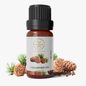 May include: A brown glass bottle of cedarwood oil with a black cap, labeled "Cedarwood Oil" and "0.33 fl. oz / 10ml." The bottle is surrounded by pine cones and sprigs of green pine needles. The label has a gold emblem.