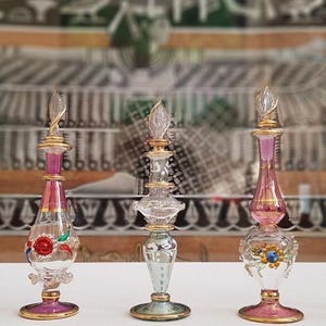 Pode incluir: Uma variedade de frascos de perfume decorativos de vidro. Os frascos são transparentes, com detalhes em rosa, dourado e azul. Cada frasco tem um design único, com tampas e bases ornamentadas. Os frascos são exibidos em uma superfície branca.