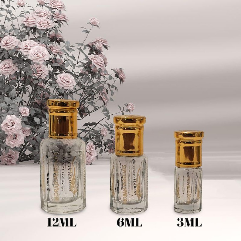 May include: Three clear glass perfume bottles with gold-colored caps, labeled 12ML, 6ML, and 3ML. The bottles have a rectangular shape with etched designs. The background features a floral arrangement of pink roses.