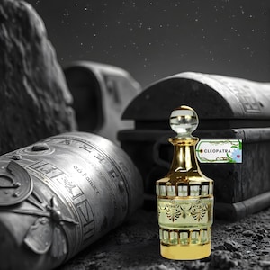 May include: A gold perfume bottle with a clear glass stopper and ornate detailing. The bottle is labelled "Cleopatra". The background includes a treasure chest and other ancient objects, all in monochrome.