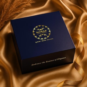 Puede incluir: Una caja azul marino con letras doradas y un logotipo dorado que dice "NORAH PERFUMES". La caja tiene las palabras "LUJO - NICHO - NATURAL" y "Abraza la Esencia de la Elegancia" impresas. La caja est&aacute; sobre una superficie de sat&eacute;n dorado.