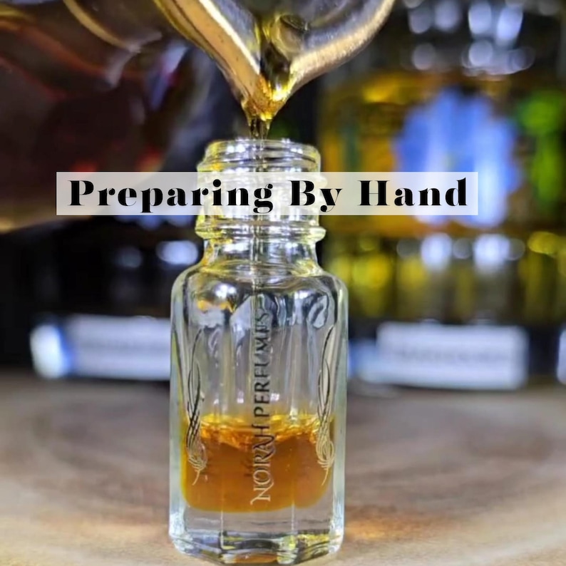 May include: A clear glass perfume bottle with a brown liquid inside is being filled with a golden liquid. The text "Preparing By Hand" is displayed above the bottle. The bottle has the words "NORAH PERFUMES" printed on it.
