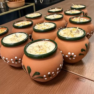 May include: A collection of terracotta clay pots filled with white wax candles. Each pot features a dark green rim and hand-painted white and green floral accents. The candle surfaces are topped with dried flower petals and a central wick.