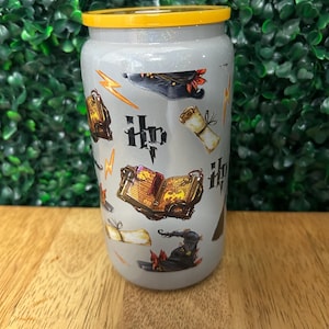 May include: A gray glitter tumbler with a yellow lid featuring a Harry Potter themed design. The design includes images of a wizard's hat, a book, a scroll, and the Harry Potter logo.