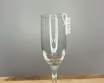 Könnte beinhalten: Eine klare Champagnerflöte aus Glas mit einem weißen Plastik-Anhänger mit der Aufschrift "BRIDE", der am Rand befestigt ist.