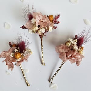 Könnte beinhalten: Drei florale Haarnadeln mit Trockenblumen in Rosa-, Burgunder- und Cremetönen. Die Blumen sind auf goldfarbenen Haarnadeln angeordnet. Der Hintergrund ist weiß mit verstreuten Blütenblattformen.