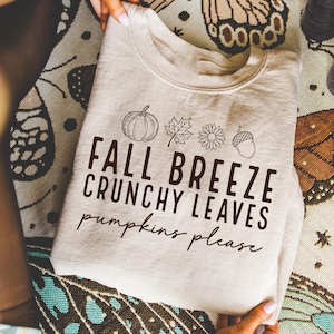 May include: A cream-colored sweatshirt with the text "FALL BREEZE CRUNCHY LEAVES pumpkins please" in a black font. The design includes a pumpkin, leaves, a flower, and an acorn. The sweatshirt is folded and held by a person.