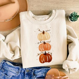 May include: A cream-coloured crewneck sweatshirt featuring a vertical watercolour illustration of three stacked pumpkins in shades of beige, light orange, and dark orange, accompanied by a tan wide-brimmed hat, blue denim jeans, and a small green succulent.