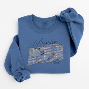 May include: A blue sweatshirt with the word "America" in script above a distressed American flag design. The number "250" is below the flag. The sweatshirt is folded on a white surface.