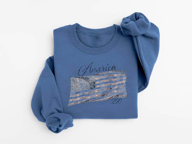 May include: A blue sweatshirt with the word "America" in script above a distressed American flag design. The number "250" is below the flag. The sweatshirt is folded on a white surface.
