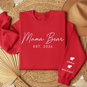 May include: A red sweatshirt with the words "Mama Bear EST. 2026" in white script. The sleeve features white bear graphics with the names "Emma" and "Michael" below. The sweatshirt is displayed on a woven surface.