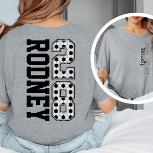 May include: A gray t-shirt with the name "Rodney" and the number "28" in a black, soccer ball-themed design on the back. The word "soccer" is printed vertically on the front of the shirt.