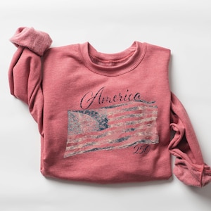 May include: A red-pink sweatshirt with the word "America" in cursive and a distressed American flag graphic. The number "250" is below the flag. The sleeves are rolled up.