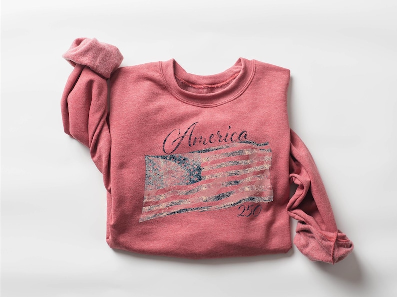 May include: A red-pink sweatshirt with the word "America" in cursive and a distressed American flag graphic. The number "250" is below the flag. The sleeves are rolled up.