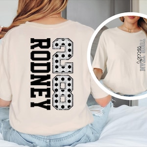 May include: Cream-colored t-shirt with the name "RODNEY" and the number "28" in black, soccer-ball-patterned font on the back. The shirt also features the text "Fall soccer" in a vertical design on the front.