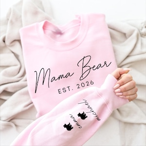 May include: A light pink sweatshirt with the words "Mama Bear EST. 2026" in black script. The sleeve features the names "Emma" and "Michael" with bear silhouettes. A cozy, casual top.