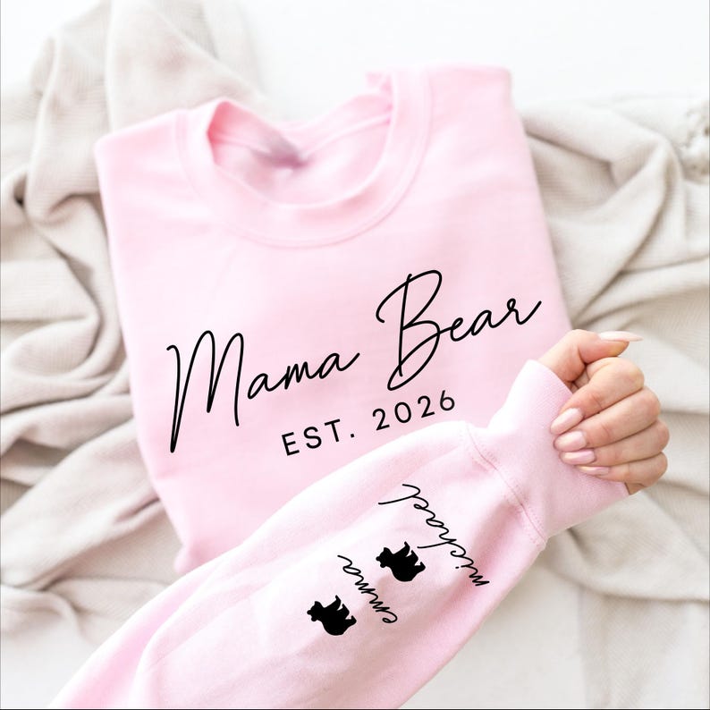May include: A light pink sweatshirt with the words "Mama Bear EST. 2026" in black script. The sleeve features the names "Emma" and "Michael" with bear silhouettes. A cozy, casual top.