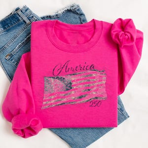 May include: A bright pink sweatshirt with the word "America" in cursive and a distressed American flag graphic. The number "250" is below the flag. The sweatshirt is laid on top of a pair of blue jeans.