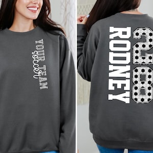 May include: Charcoal gray sweatshirt with the text "YOUR TEAM soccer" vertically on the front. The back features the name "RODNEY" and the number "28" in a soccer ball design. A comfortable, casual top for soccer fans.