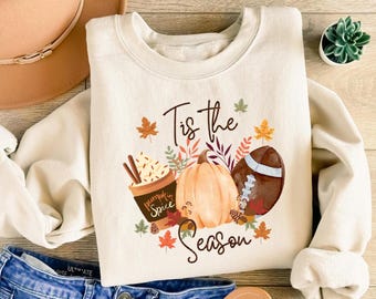 May include: Cream-colored sweatshirt with a fall-themed graphic. The design includes a pumpkin spice latte, a pumpkin, and a football, with the text "Tis the Season". Autumn leaves and accents complete the design.