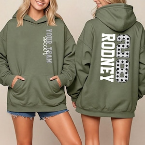 May include: Olive green hooded sweatshirt with front pocket and drawstring. The front features the text "YOUR TEAM soccer" in a vertical design. The back displays the name "RODNEY" and the number "28" with soccer ball graphics.