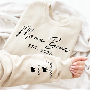 May include: A beige sweatshirt with the words "Mama Bear" and "EST. 2026" in black script. The sleeve features two black bear silhouettes and the names "Emma" and "Michael".