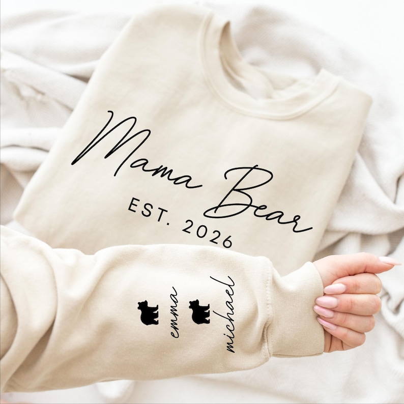 May include: A beige sweatshirt with the words "Mama Bear" and "EST. 2026" in black script. The sleeve features two black bear silhouettes and the names "Emma" and "Michael".