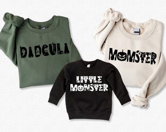 Peut inclure: Trois sweats à capuche sur le thème d'Halloween en vert, beige et noir. Le sweat vert dit "DADCULA" en lettres noires avec un fantôme, une pierre tombale et d'autres icônes d'Halloween. Le sweat beige dit "MOMSTER" en lettres noires avec une citrouille, un fantôme et d'autres icônes d'Halloween. Le sweat noir dit "LITTLE MONSTER" en lettres blanches avec une citrouille, un fantôme et d'autres icônes d'Halloween.