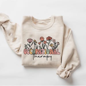 May include: A beige sweatshirt with the text "Occupational Therapy" in colorful letters with a floral design below.
