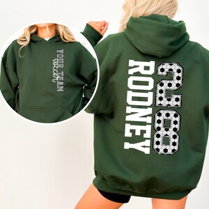 May include: Dark green hooded sweatshirt with white text on the back that reads "RODNEY 2888". The front of the sweatshirt has the text "YOUR TEAM soccer" in white. The design incorporates soccer ball patterns.