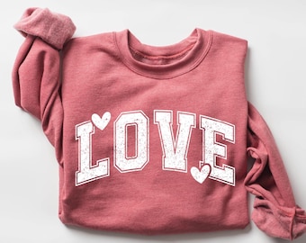 May include: A dusty rose sweatshirt with the word "LOVE" in large, distressed white letters. Two small white hearts are placed above and below the "O" and "E". The sleeves are cuffed, and the background is white.