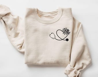 May include: A cream-colored sweatshirt featuring a black stethoscope design shaped like a heart, with floral accents. The design includes a flower and a small black flower. The sleeves are rolled up.