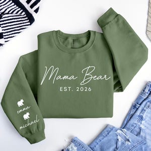 May include: An olive green sweatshirt with the words "Mama Bear" and "EST. 2026" in white script. The left sleeve features two white bear silhouettes with the names "emma" and "michael" below. The sweatshirt is folded on a white surface.