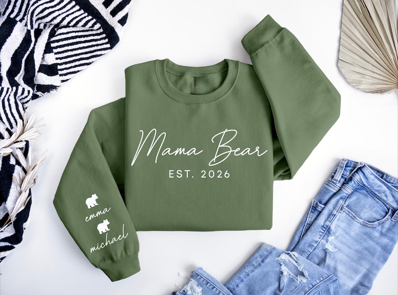 May include: An olive green sweatshirt with the words "Mama Bear" and "EST. 2026" in white script. The left sleeve features two white bear silhouettes with the names "emma" and "michael" below. The sweatshirt is folded on a white surface.