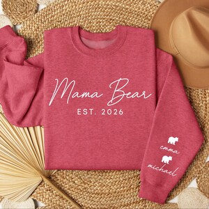 May include: A red sweatshirt with the words "Mama Bear EST. 2026" in white script. The sleeve features white bear graphics with the names "Emma" and "Michael" below. The sweatshirt is displayed on a woven surface with a hat.
