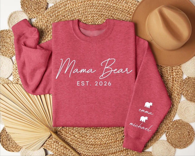 May include: A red sweatshirt with the words "Mama Bear EST. 2026" in white script. The sleeve features white bear graphics with the names "Emma" and "Michael" below. The sweatshirt is displayed on a woven surface with a hat.