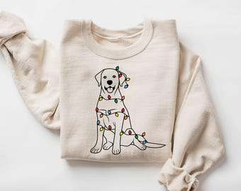 May include: Cream-colored sweatshirt featuring a black outline illustration of a Labrador Retriever dog adorned with colorful Christmas lights. The dog is sitting with its tongue out. The sweatshirt has long sleeves and a crew neck.