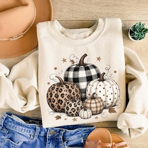 May include: A cream-colored crewneck sweatshirt featuring a graphic of five pumpkins with various patterns including black and white buffalo plaid, leopard print, and polka dots, accented by small brown stars and swirling vine illustrations.