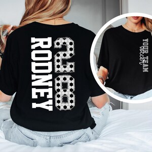 May include: Black t-shirt with the name "RODNEY" and the number "28" in white, soccer ball-themed design. The shirt also features the text "YOUR TEAM soccer" in white. The shirt is a casual style.