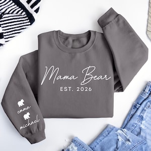 May include: A gray sweatshirt with the words "Mama Bear" and "EST. 2026" in white script. The left sleeve has two white bear graphics with the names "emma" and "michael" below. The sweatshirt is paired with blue jeans.