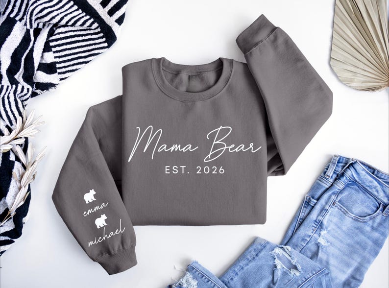 May include: A gray sweatshirt with the words "Mama Bear" and "EST. 2026" in white script. The left sleeve has two white bear graphics with the names "emma" and "michael" below. The sweatshirt is paired with blue jeans.