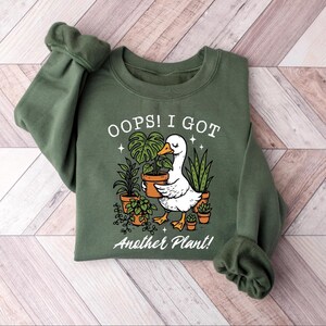 May include: Green sweatshirt with the text "OOPS! I GOT Another Plant!" and a graphic of a white goose holding a potted plant. The goose is surrounded by other potted plants in terracotta pots. The sweatshirt has long sleeves.
