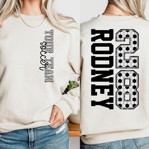 May include: Cream-colored sweatshirt with the text "YOUR TEAM soccer" in black on the front. The back features the name "RODNEY" and the number "28" in black, with a soccer ball pattern.